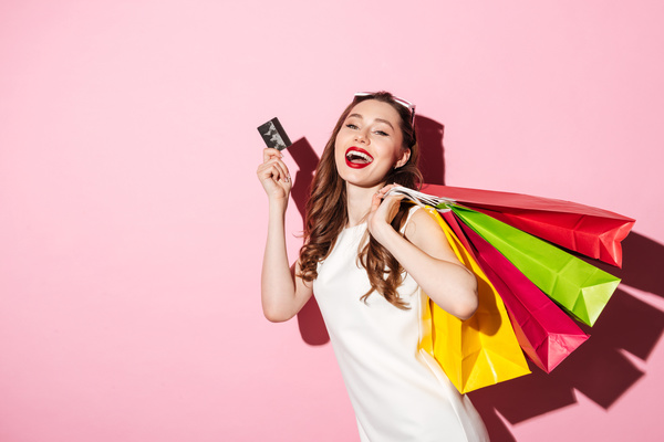 Picture of a cheerful young brunette woman in white summer dress holding credit card posing with shopping bags and looking at camera over pink background.